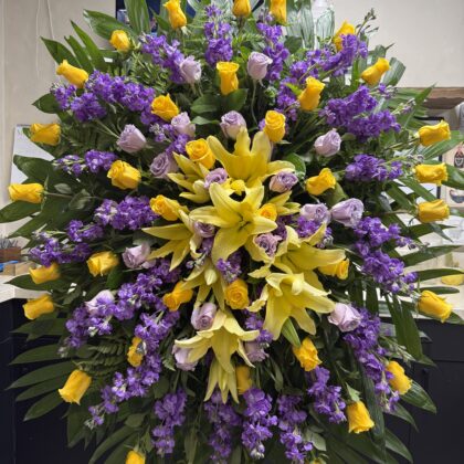 Whispering Pines Funeral Spray - Celestial Elegance featuring yellow lilies, roses, and purple stock flowers on a stand.