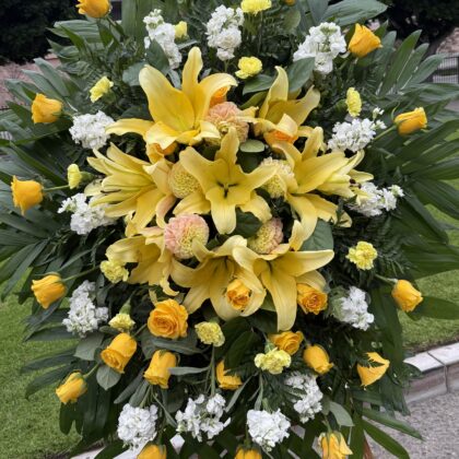 Bright Sacred Tribute Funeral Spray Arrangement featuring yellow lilies, roses, and white stock flowers on palm leaves.