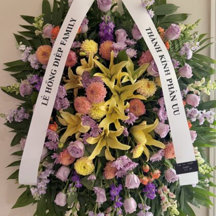 Joyful Remembrance Funeral Spray for Celebrating Life featuring yellow lilies, lavender roses, and dahlias on palm leaves.
