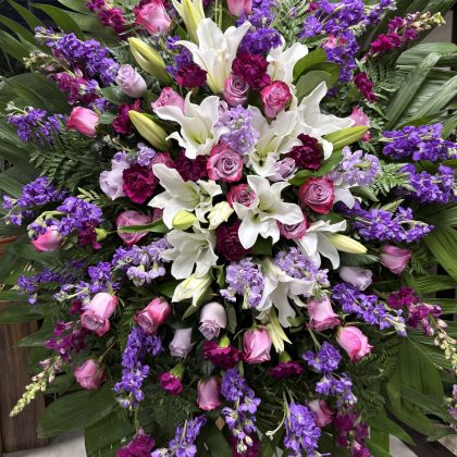 Exquisite Meadow of Memories Funeral Spray with white lilies, purple stock, and lavender roses on palm leaves.