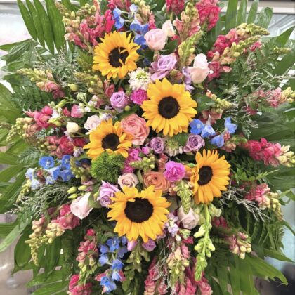 A Vibrant Rainbow Bridge Funeral Spray featuring sunflowers, roses, and snapdragons in a lush, elegant arrangement.