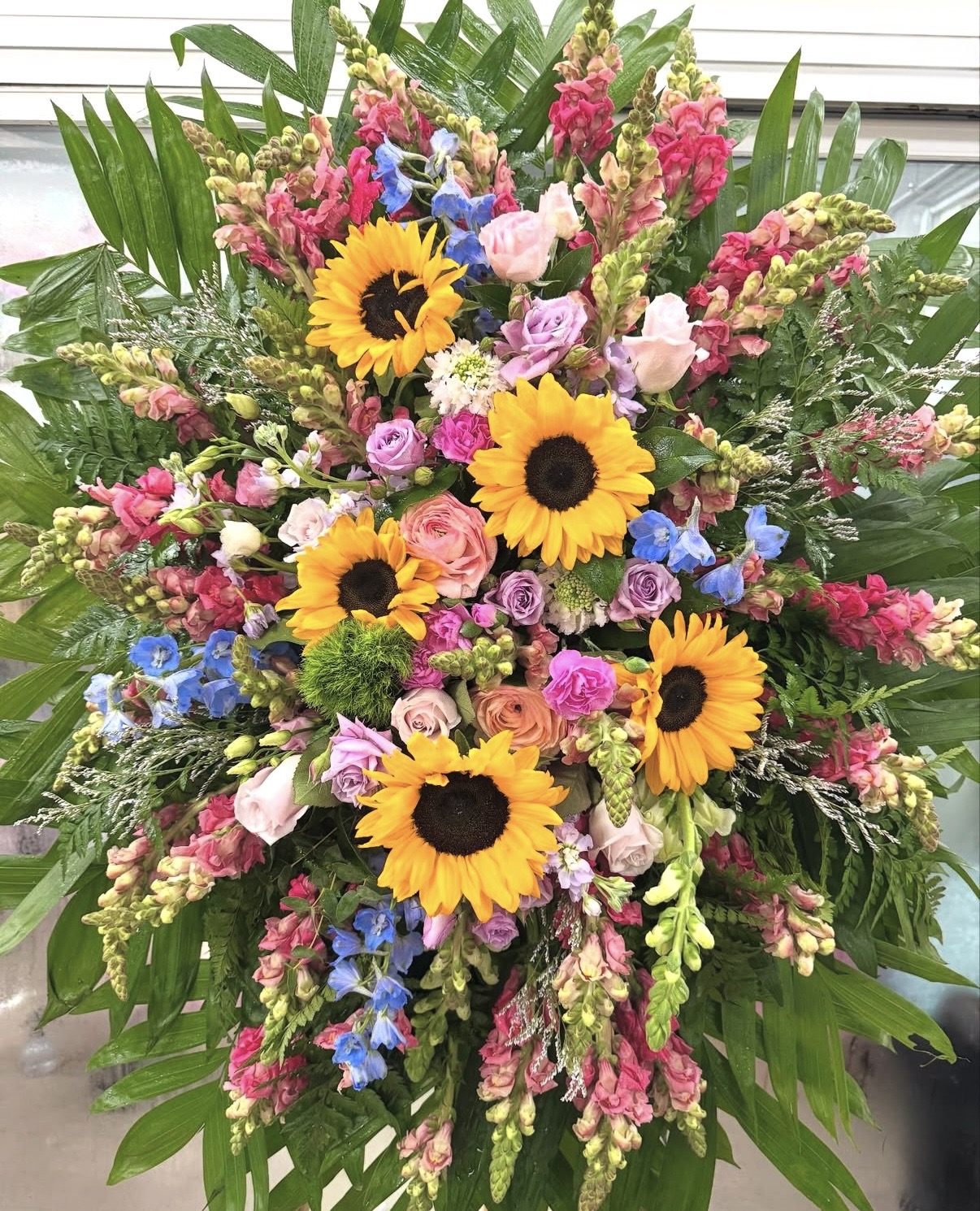 A Vibrant Rainbow Bridge Funeral Spray featuring sunflowers, roses, and snapdragons in a lush, elegant arrangement.