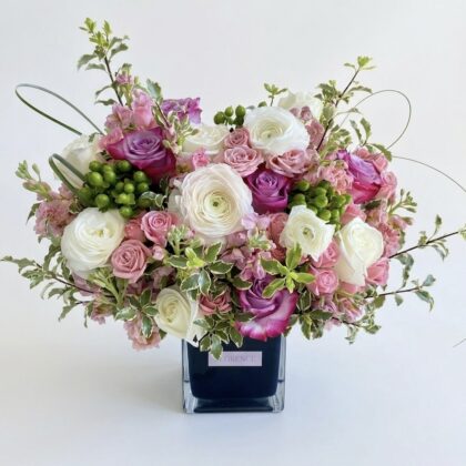 Prato Di Fiore - Wood Pot features a luxurious arrangement of pink and purple roses, white ranunculus, and delicate greenery.