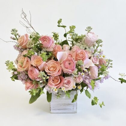 Luxurious arrangement of soft pink roses, delicate lilacs, and lush greenery in a chic white container.