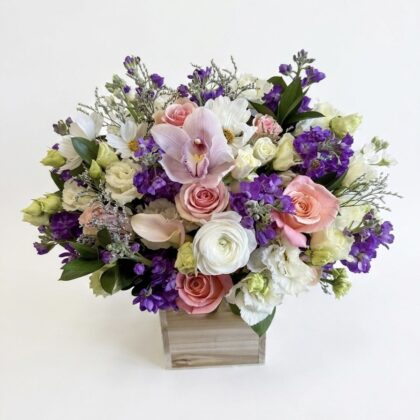 Luxurious floral arrangement featuring pink roses, white orchids, and purple blooms in a refined wooden box.