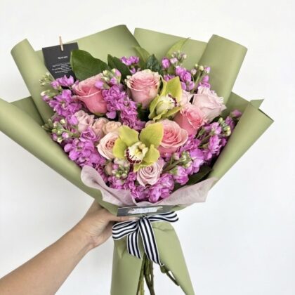 Elegant bouquet featuring pink roses, purple stock, and green orchids, beautifully arranged with lush foliage.
