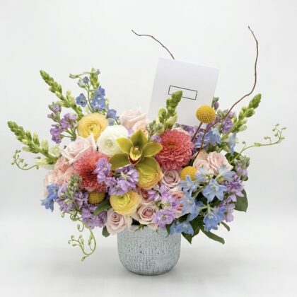 Luxurious arrangement featuring soft pink roses, vibrant dahlias, and delicate blue delphiniums in a textured vase.
