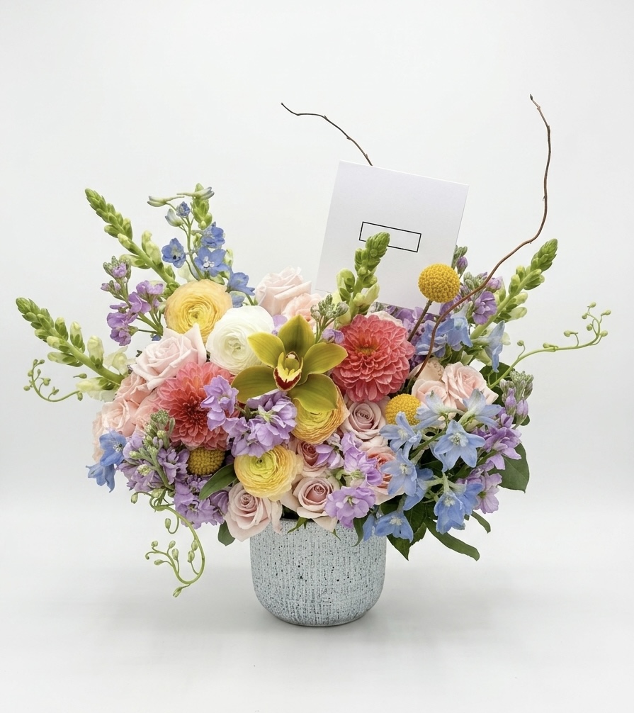 Luxurious arrangement featuring soft pink roses, vibrant dahlias, and delicate blue delphiniums in a textured vase.