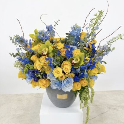 Mykonos – Striking Concrete Vase filled with a luxurious arrangement of yellow roses, blue delphiniums, and green orchids.