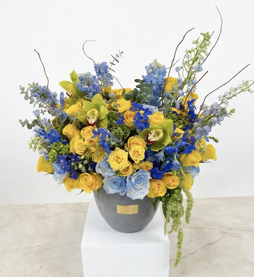 Mykonos – Striking Concrete Vase filled with a luxurious arrangement of yellow roses, blue delphiniums, and green orchids.