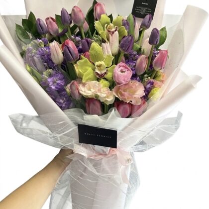 A hand holds a Maison De Seine wrapped bouquet featuring a Vibrant Tulip Bouquet with orchids and purple accents.