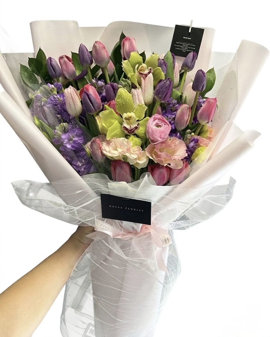 A hand holds a Maison De Seine wrapped bouquet featuring a Vibrant Tulip Bouquet with orchids and purple accents.
