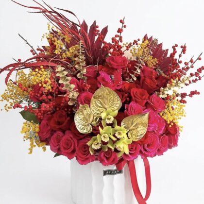 New Beginnings – Opulent Floral Design featuring red roses, gold anthuriums, and yellow mimosa in a white ceramic vase.
