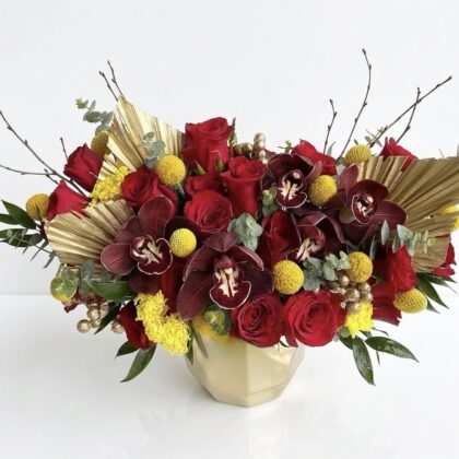 Regal Sweet Prosperity Ceramic Vase featuring deep red roses, dark orchids, yellow billy balls, and gold palm leaves.