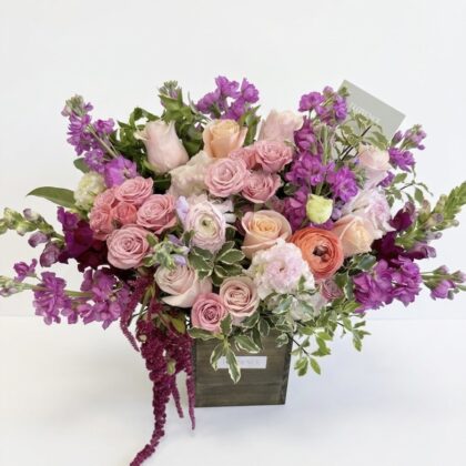 Exquisite Bebe Macaron Wood Pot Arrangement featuring soft pink roses and vibrant purple stock flowers in a rustic box.