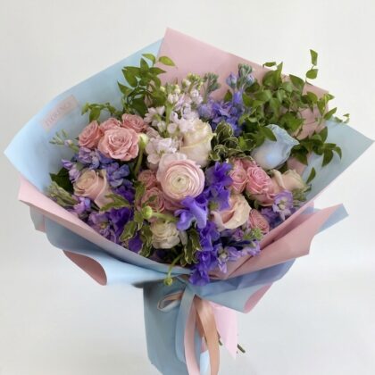 Whimsical Bouquet De Cherie Wrapped Bouquet featuring soft pink roses, purple delphiniums, and lush greenery.