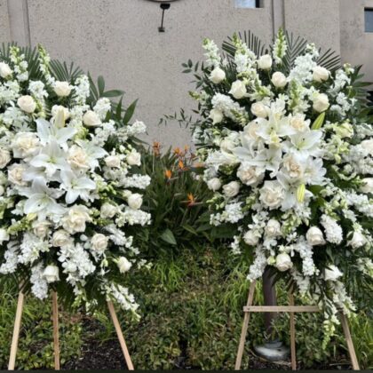 Two Heritage Classic White Funeral Spray arrangements featuring lilies, roses, and snapdragons on wooden stands.