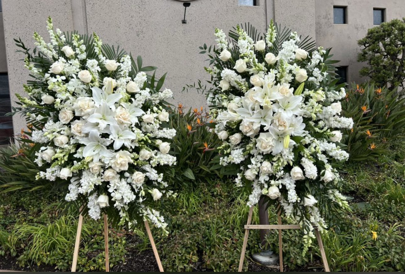 Two Heritage Classic White Funeral Spray arrangements featuring lilies, roses, and snapdragons on wooden stands.