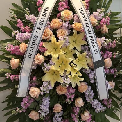 Sweet Melody Funeral Spray with Yellow Lilies, featuring soft peach roses and lavender stock flowers on a wooden easel.