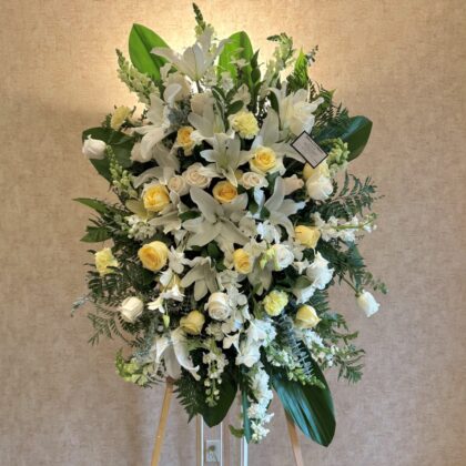 Bright Memories – Elegant Funeral Spray featuring white lilies, pale yellow roses, and lush greenery on a wooden stand.