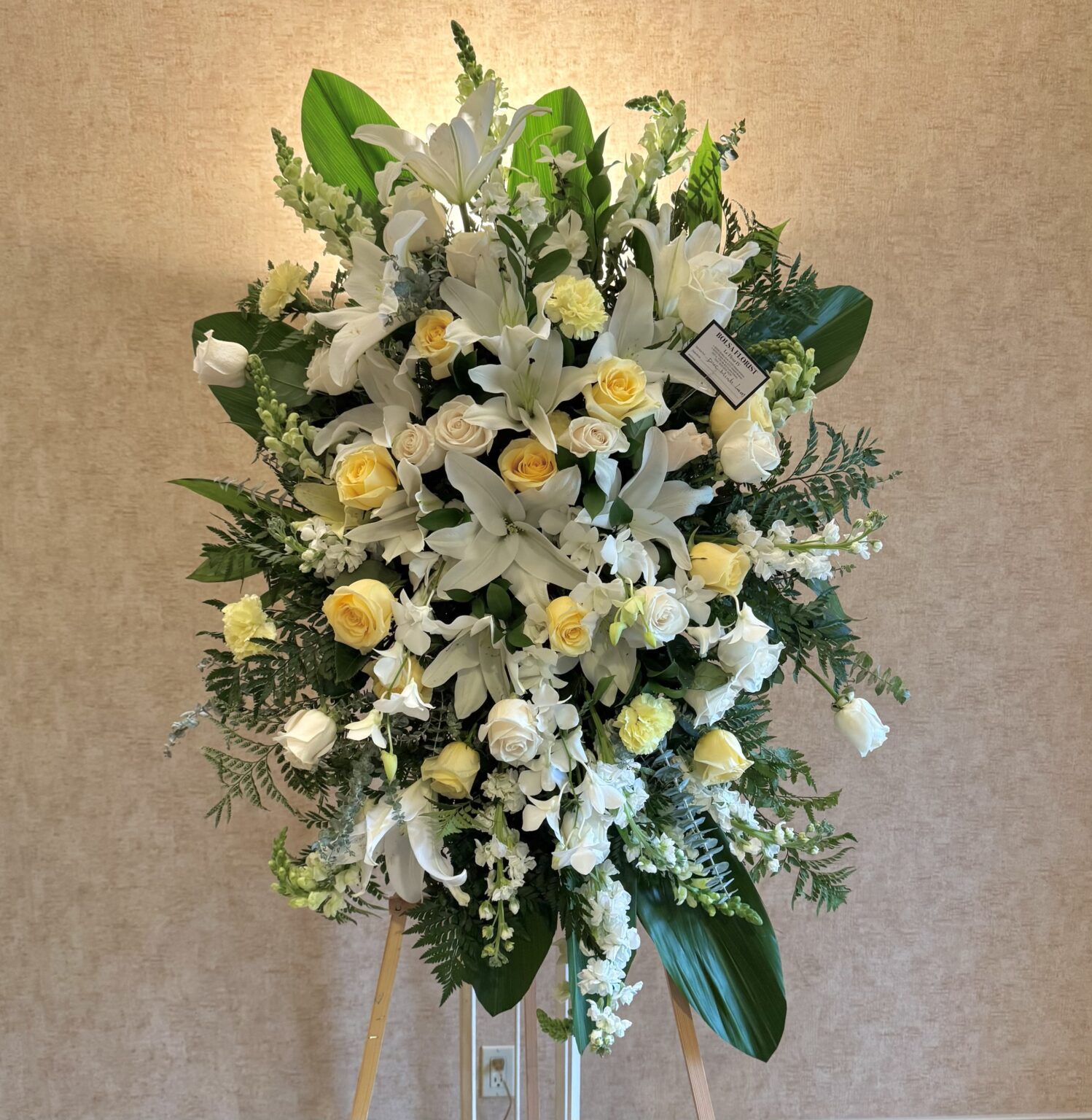 Bright Memories – Elegant Funeral Spray featuring white lilies, pale yellow roses, and lush greenery on a wooden stand.