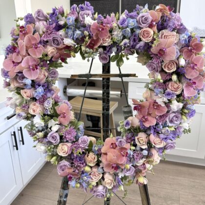 Sweetest Solace &ndash; Heart Wreath for Farewell featuring mauve roses, pink orchids, and purple accents on a display stand.