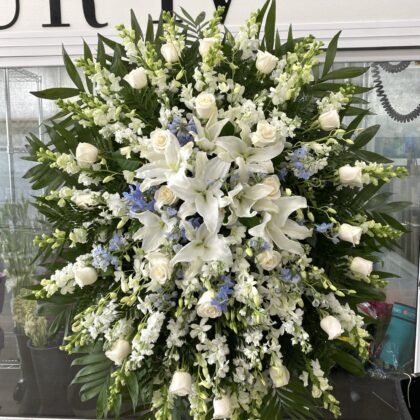 Forever Loved Funeral Spray - Serene Floral Arrangement featuring white lilies, roses, and blue delphinium on a stand.
