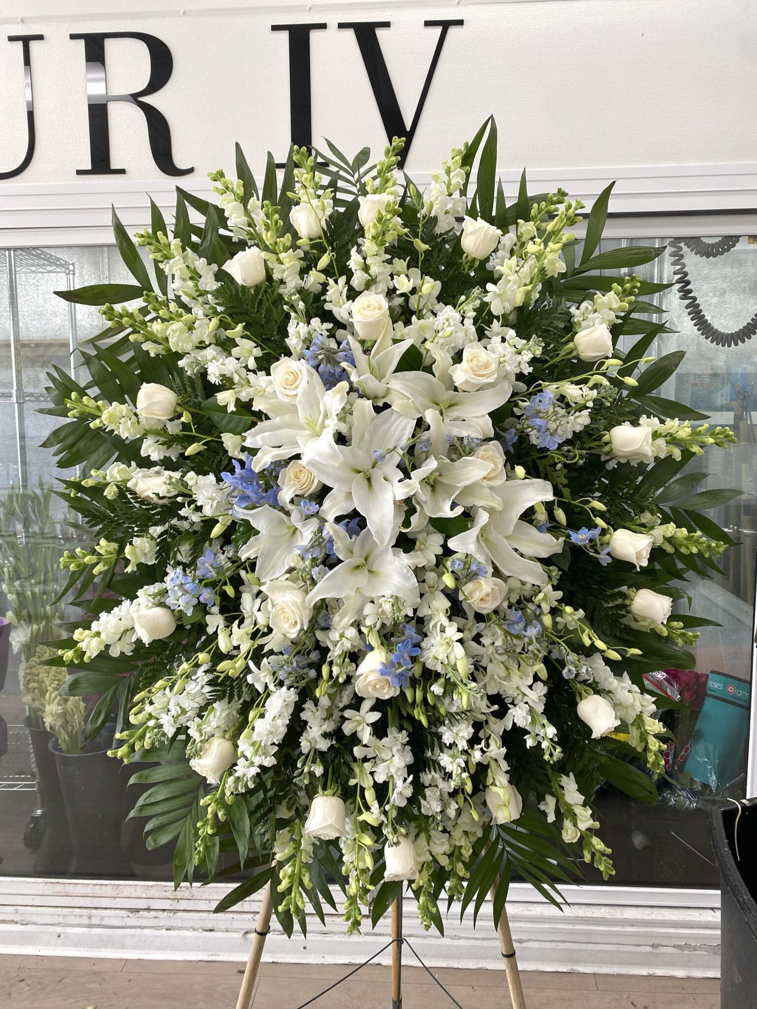 Forever Loved Funeral Spray - Serene Floral Arrangement featuring white lilies, roses, and blue delphinium on a stand.