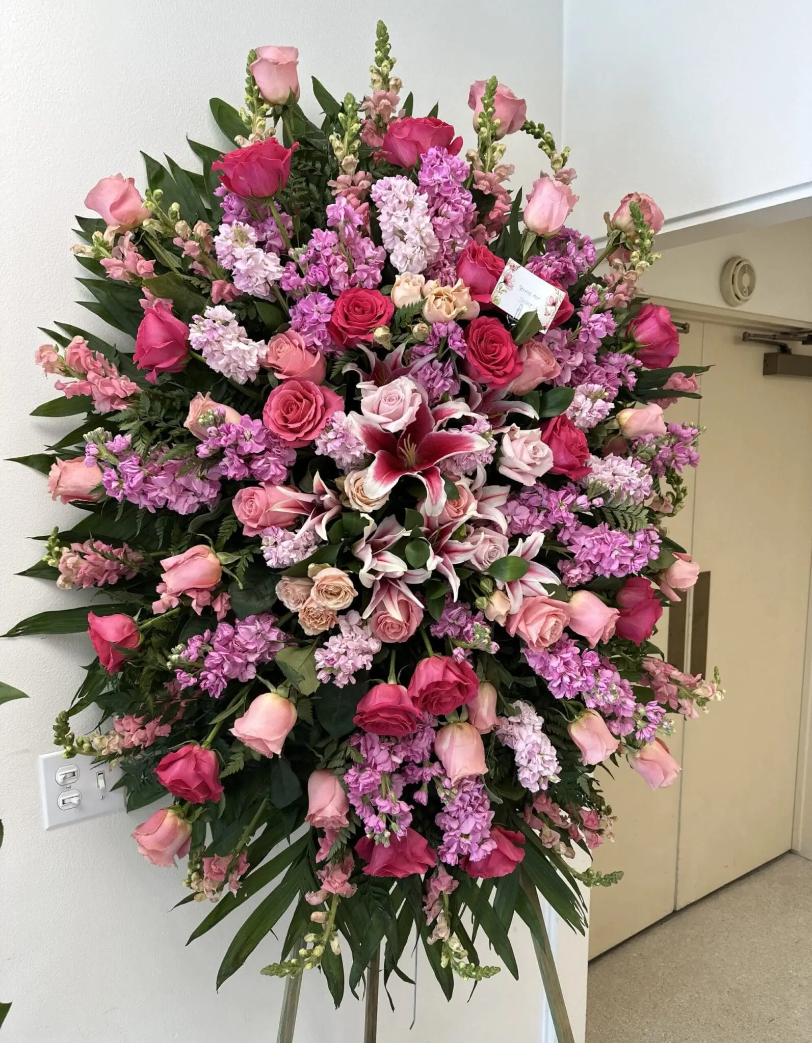 Sweet Serenity Funeral Spray – Lush Floral Arrangement featuring pink roses, lilies, and stock on a standing easel.