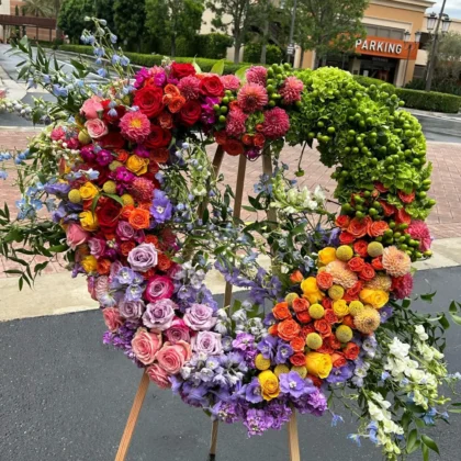 Rainbow Remembrance Heart Wreath – Colorful Tribute featuring vibrant roses, dahlias, and hydrangea on a wooden stand.