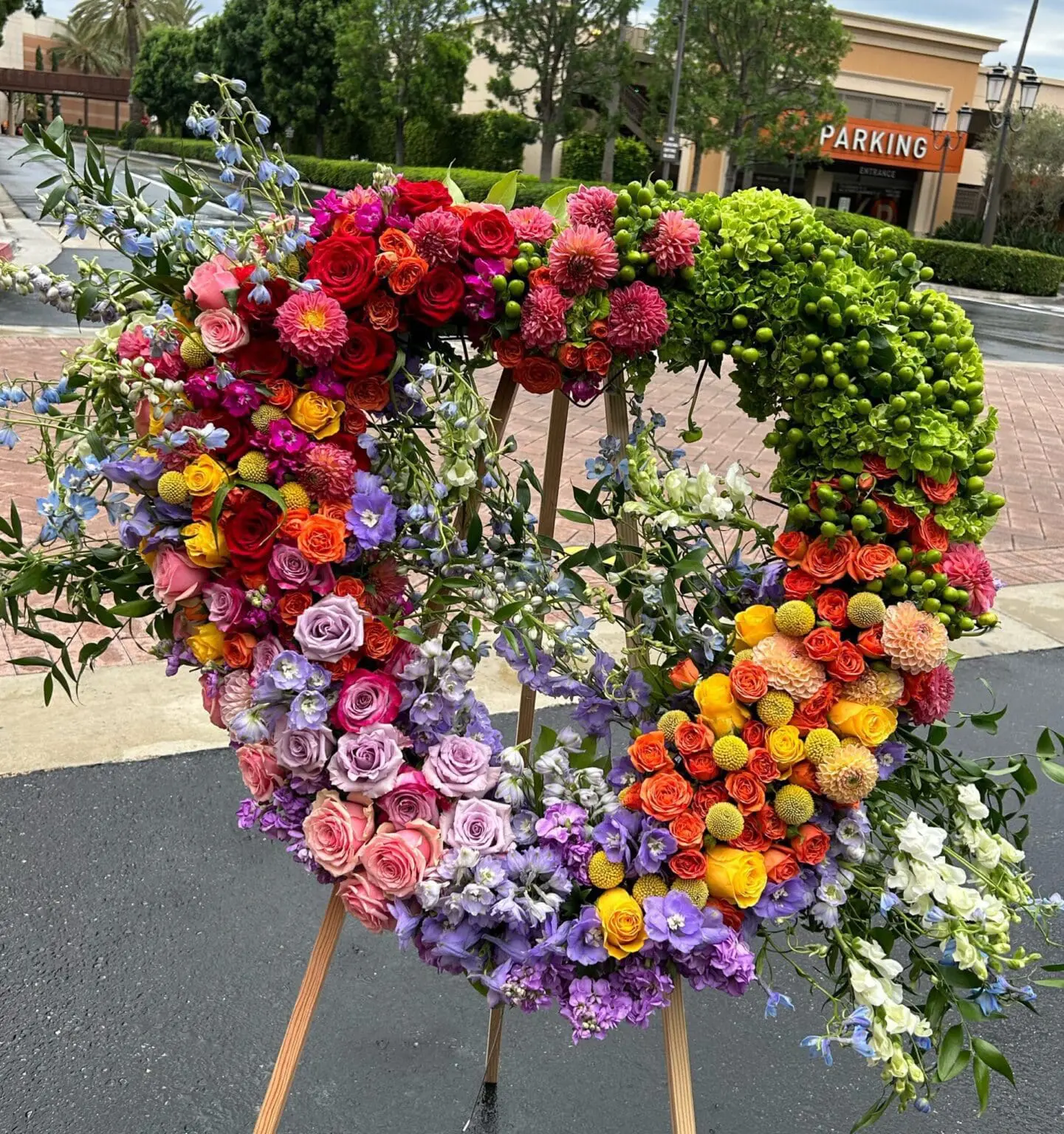 Rainbow Remembrance Heart Wreath – Colorful Tribute featuring vibrant roses, dahlias, and hydrangea on a wooden stand.