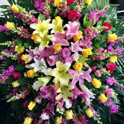 Feathers & Flora Funeral Spray – Vibrant Celebration featuring yellow lilies, pink snapdragons, and roses in lush greenery.