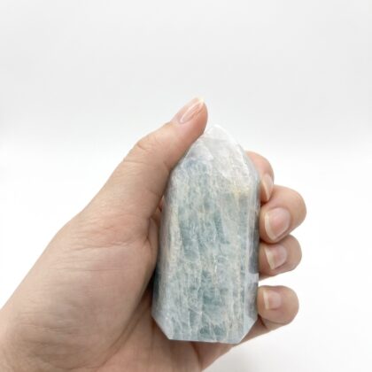 A hand holds a polished, hexagonal aquamarine crystal tower with pale blue and white veined textures.