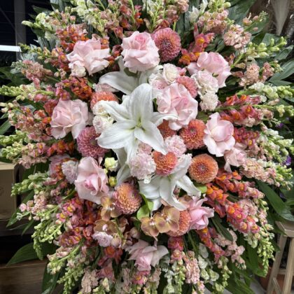 A luxurious funeral standing spray featuring soft pink roses, white lilies, and peach dahlias arranged with lush greenery.