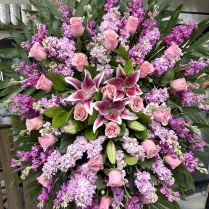 A grand funeral spray featuring pink roses, purple stock, and speckled lilies arranged with lush green palm leaves.
