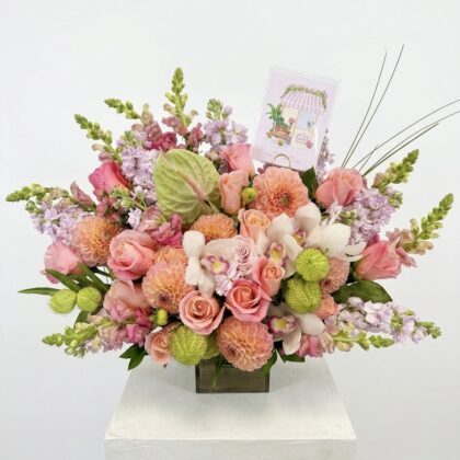 A luxurious floral arrangement featuring peach roses, dahlias, and lavender snapdragons in a glass vase.