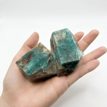 A hand holds a raw, dual-pointed teal and brown crystal specimen against a clean white background.