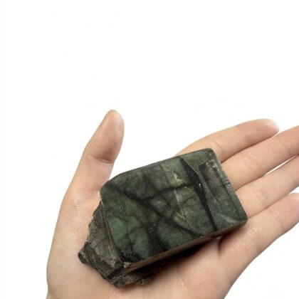 A hand holds a rectangular, dark green emerald specimen with intricate black veining and a raw, textured base.
