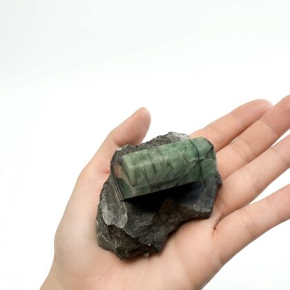 A hand holds a raw, rectangular emerald crystal embedded in a dark, jagged piece of natural matrix rock.