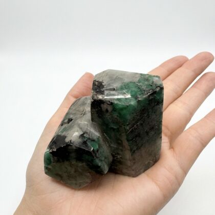 A hand holds two polished, hexagonal emerald crystals featuring deep forest green hues and dark mineral inclusions.