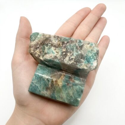 A hand holds a raw, rectangular piece of teal and brown apatite crystal against a clean white background.