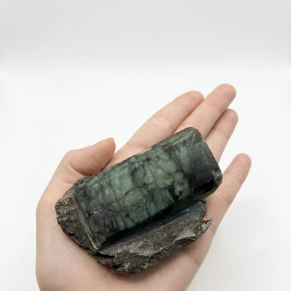 A hand holds a raw, rectangular emerald crystal with deep forest green hues and natural veining on a dark matrix.