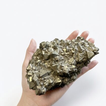 A hand holds a cluster of metallic pyrite crystals with sharp, geometric, gold-toned facets against a white background.