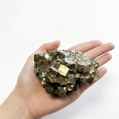 A hand holds a raw, metallic pyrite cluster with shimmering, golden cubic crystals against a clean white background.