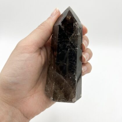 A hand holding a polished, dark brown Grounding Smoky Quartz Point for Spiritual Uplift against a white background.