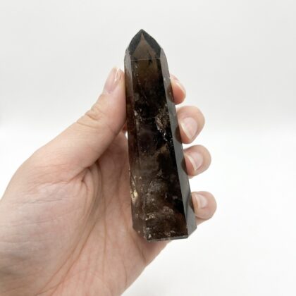 A hand holds a polished, dark brown Grounding Smoky Quartz Point for Spiritual Uplift against a white background.