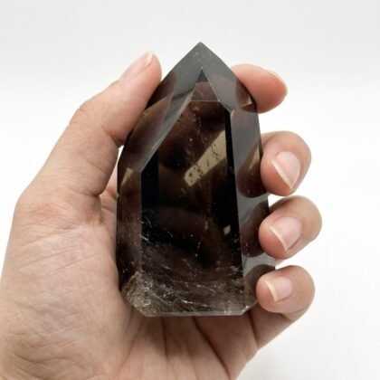 A hand holds a polished, dark brown Grounding Smoky Quartz Point for Spiritual Uplift against a white background.