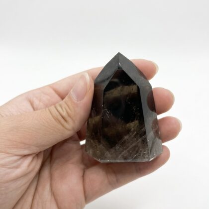 A hand holds a polished, dark Smoky Quartz &ndash; Grounding Crystal Point against a clean, white background.