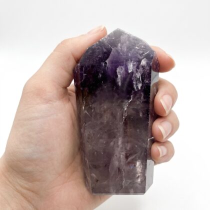 A hand holds a polished Amethyst &ndash; Beautiful Crystal Tower Keepsake with deep purple hues against a white background.
