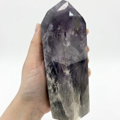 A hand holding a polished Amethyst – Stunning Crystal Tower Gift with deep purple and smoky grey natural inclusions.